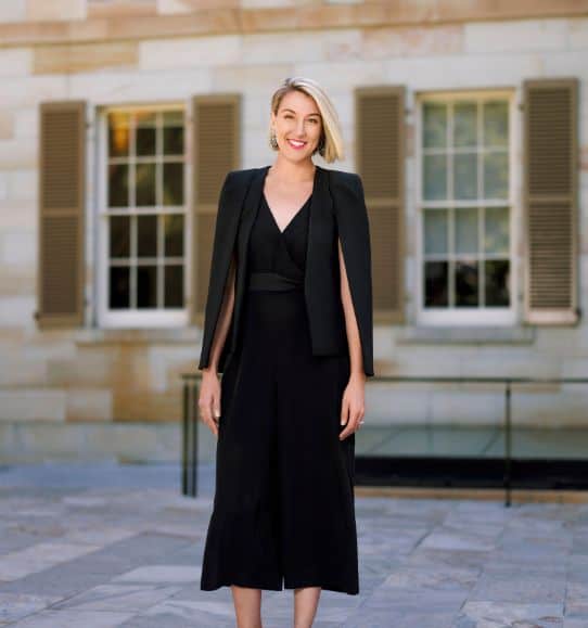 Roxy Topham, wearing a black jumpsuit, stands in front of a building.
